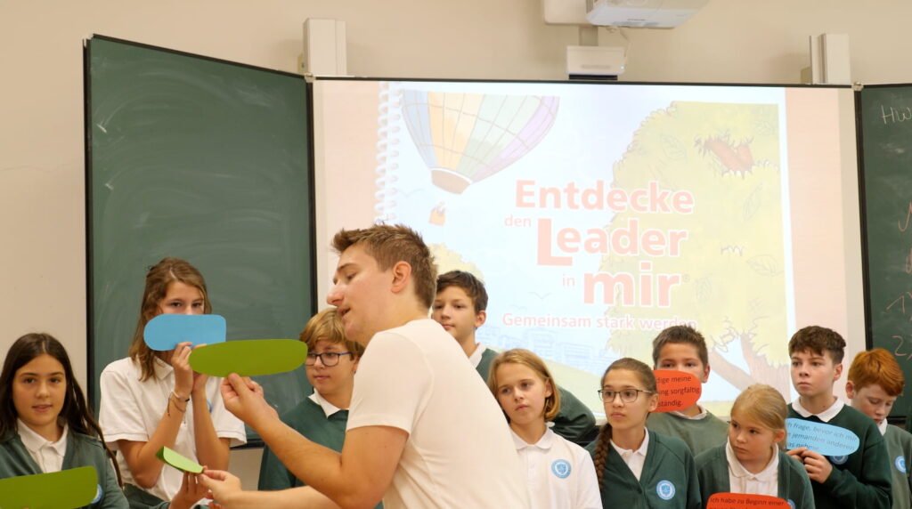 Ein Lehrer arbeitet mit einer Schulklasse, im Hintergrund die Projektion „Entdecke den Leader in mir“