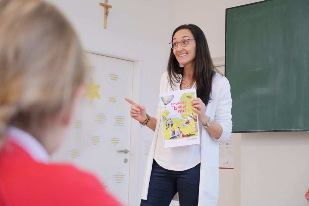 Eine Lehrerin zeigt lachend das Buch „Entdecke den Leader in mir“ vor ihrer Klasse
